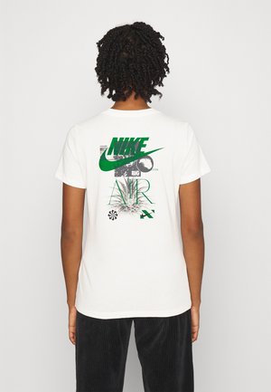 Osoba o kręconych włosach nosi białą koszulkę Nike z zielonym znakiem i abstrakcyjną zielono-czarną grafiką na plecach.