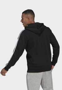 Hoodie preto com cordão e riscas brancas nos ombros, combinado com calças de fato cinzentas. O material parece suave e texturizado.