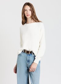 Junge Frau mit langen Haaren, die einen weißen Rippensweater trägt, der in eine hochtaillierte hellblaue Jeans mit einem dunkelbraunen Gürtel gesteckt ist.