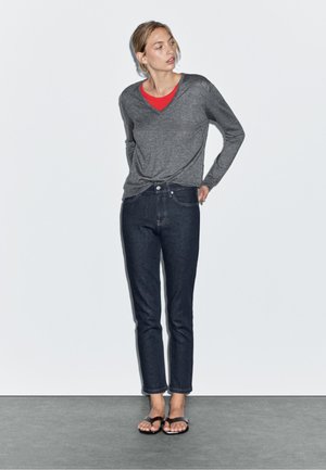 Grå långärmad tröja över en röd T-shirt, kombinerad med mörkblå jeans och svarta flip-flops; slät tyg och figurnära snitt. Minimalistisk bakgrund.