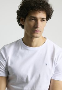 Camiseta de algodón blanca con cuello redondo, mangas cortas y un pequeño logo azul marino en el pecho. Textura suave y un corte ajustado.