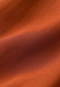 Lähikuva pehmeästä, ruosteenvärisestä kankaasta, jossa on hieno rakenne ja lempeät taitteet, korostaen materiaalin sileää ja hieman teksturoidumpaa pintaa.