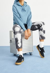 Felpa azzurra con cappuccio e cordino, pantaloni tie-dye grigi e bianchi, sneakers nere con accenti bianchi e calzini bianchi. Seduto su una valigia argentata.