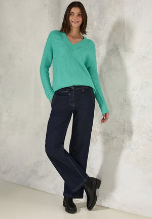 Teal gestreifter Pullover mit V-Ausschnitt, kombiniert mit dunkelblaue Schlagjeans. Schwarze Stiefeletten runden das Outfit vor einem strukturierten Hintergrund ab.