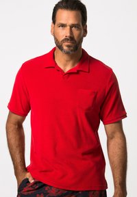 Rotes Poloshirt mit kurzen Ärmeln, einer linken Brusttasche und einem klassischen Kragen. Weicher Stoff. Getragen mit gemusterten Shorts.