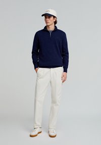 Marineblå strikpullover med lynlås krave, cremefarvede plisserede bukser og hvide sneakers med brune såler. Modellen bærer en lysefarvet kasket.