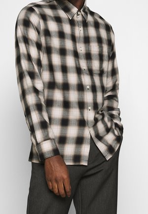 Mann trägt ein schwarz-weiß-beiges kariertes Hemd mit Knöpfen und Brusttasche sowie dunkelgraue Hose, eine Hand in der Tasche.