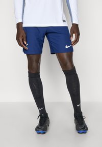 Marinesport shorts med teksturerede striber, hvidt Nike-logo, parret med sorte fodboldsokker og sorte støvler med blå accenter.