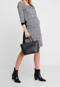 Svart strukturerad handväska med dubbla handtag, ficka med dragkedja och guldfärgade detaljer. Modellen bär en grå rutig klänning med trekvartslånga ärmar och svarta accenter.