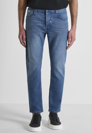 Hombre con vaqueros ajustados azul y zapatillas negras con suelas blancas, de pie contra un fondo claro y liso.