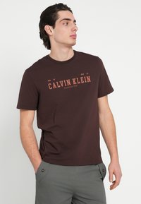 Smeđa pamučna majica s tiskanim logotipom "CALVIN KLEIN" u narančastoj boji, okruglim izrezom, kratkim rukavima i opuštenim krojem.