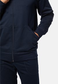 Granatowy hoodie na zamek błyskawiczny z kieszeniami typu kangur, ściągaczami na rękawach i miękką fakturą. Luźniejszy krój z subtelnymi detalami szycia, w zestawie z dopasowanymi spodniami.