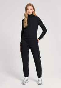 Giacca nera con zip e collo alto abbinata a pantaloni cargo; i pantaloni hanno tasche laterali, orli stretti e una texture di tessuto liscia.