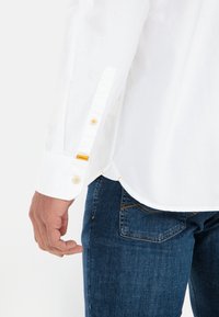 Witte longsleeve shirt met een knoopmanchet en contrasterende gele knoopaccenten, gecombineerd met donkerblauwe spijkerbroek.