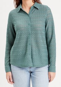 Chemise boutonnée verte texturée avec col, manches longues et motif ondulé. Comprend des boutons assortis et une coupe décontractée.