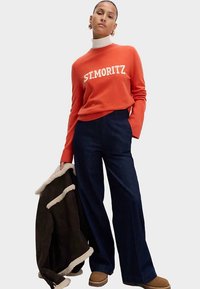 Pull rouge col rond avec le texte « ST. MORITZ », associé à un pantalon large et sombre. Le mannequin tient une veste marron doublée en shearling et porte des bottes beiges.