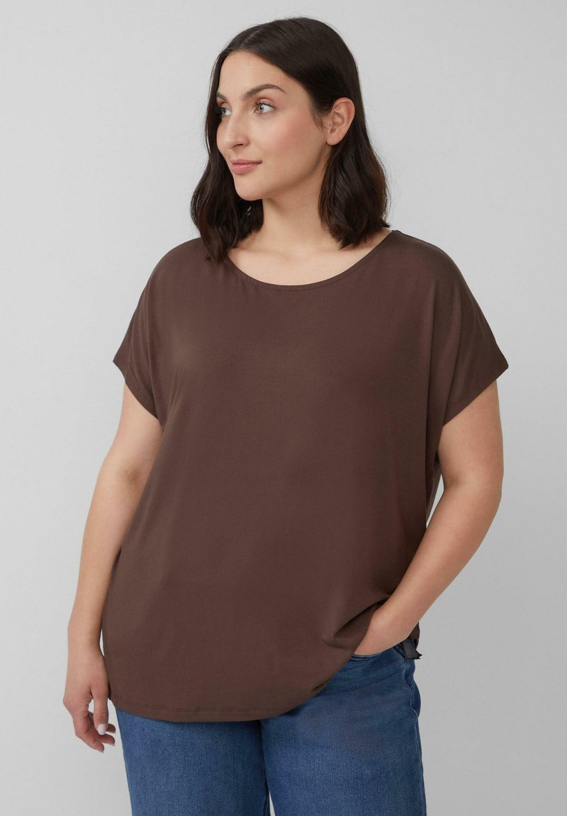 Vrouw met schouderlang donker haar, gekleed in een losse bruine top met korte mouwen en blauwe jeans, staande met één hand in haar zak en kijkend naar links.