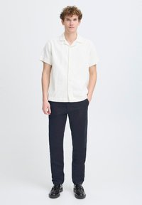 Chemise blanche à manches courtes et texture, associée à un pantalon noir ajusté et des chaussures en cuir noir. Design simple, lignes épurées.
