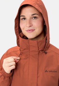 Jonge vrouw die het trekkoord van de capuchon van een roestkleurige Vaude-jack aanpast, lachend met een neutrale witte achtergrond.