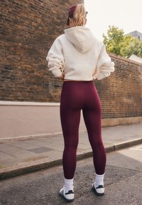 Person steht auf der Straße, trägt einen weißen Kapuzenpullover, weinrote Leggings, weiße Socken, Turnschuhe und eine weinrote Mütze, blickt auf eine Backsteinmauer.