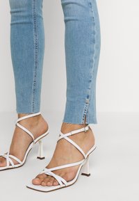 Nohy obuté v bílých sandálech na vysokém podpatku s popruhy, které ladí se světle modrými úzkými džíny s malým bočním rozparkem a třepícími se lemy.