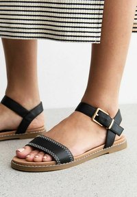 Sandalias de cuero negro con una banda ancha, costuras blancas y una correa para el tobillo con un hebilla dorada. Suela marrón y textura suave.