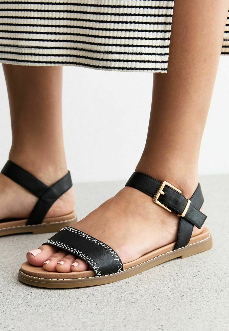 Sandalias de cuero negro con una banda ancha, costuras blancas y una correa para el tobillo con un hebilla dorada. Suela marrón y textura suave.