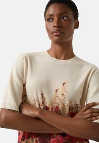 Kremowy, prążkowany sweter dzianinowy z krótkim rękawem, zdobiony kwiatowym wzorem w odcieniach czerwieni i pomarańczy na dolnej połowie. Gładka faktura.