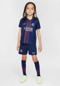 Kind steht in einem dunkelblauen Paris Saint-Germain Fußballtrikot mit Sponsorlogo, passenden Socken und hellen Schuhen.