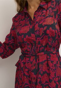 Donna che indossa un vestito nero con bottoni, maniche lunghe e stampa floreale rossa brillante, cintrata in vita, su uno sfondo neutro.