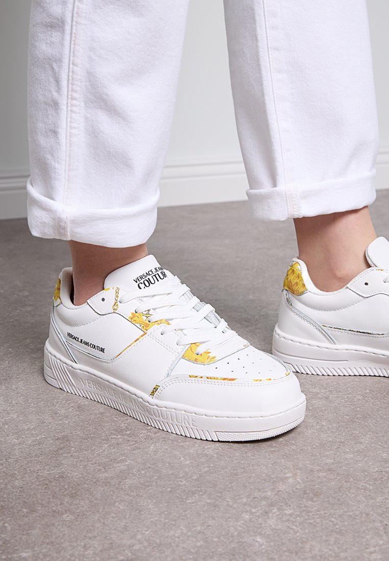 Zapatillas blancas con acentos florales amarillos, suela de goma texturizada, puntera perforada y con la marca "VERSACE JEANS COUTURE" en el lado.