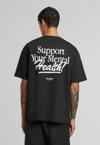 Sort bomulds t-shirt med fed hvid tekst på ryggen: "Støt dit mentale helbred!" med et logo nær kanten. Korte ærmer, afslappet pasform.