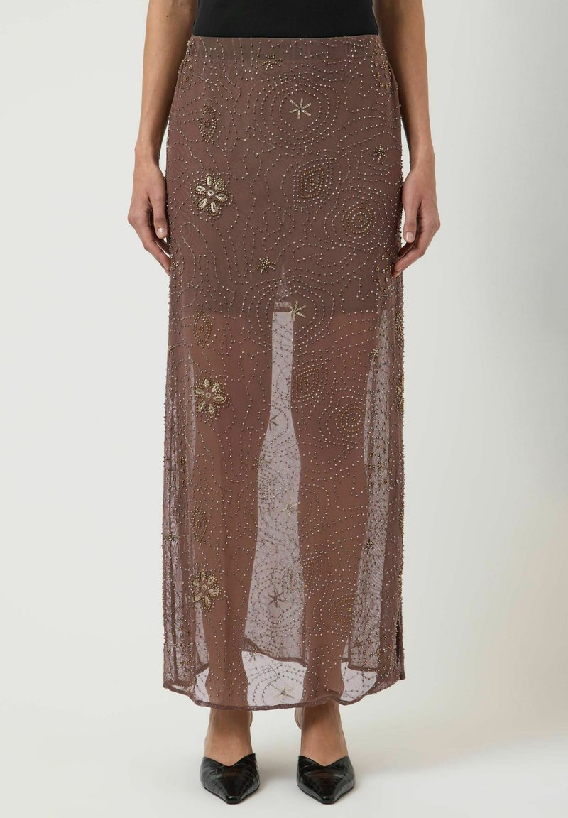 Jupe maxi marron transparente avec broderie florale et en spirale de perles dorées portée avec des chaussures noires pointues et un top.