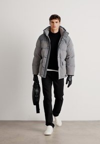 Chaqueta de plumas gris aislante con cuello alto y cierre de botones, combinada con un suéter negro, pantalones negros y zapatillas blancas.