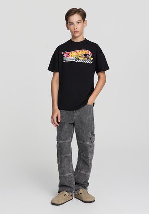 T-shirt nera in cotone con una grafica colorata di Hot Wheels, abbinata a pantaloni cargo grigi e scarpe slip-on beige. Il modello sta in piedi diritto.