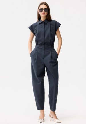 Kvinde med langt brunt hår iført sort kortærmet tekstureret jumpsuit, sorte solbriller og hvide slingback-hæle, stående med hænderne i lommerne.