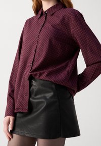 Camisa burdeos de botón con un patrón geométrico, con mangas largas y cuello, combinada con una minifalda de cuero negra.
