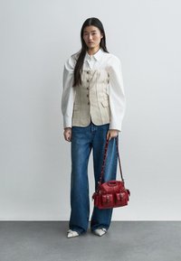 Nő fehér, puffos ujjú ingben, bézs gombos fűzővel, bőszárú farmerral, fehér hegyes orrú cipővel, piros bőr kézitáskával a kezében.