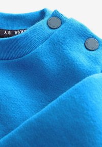 Suéter azul brillante con cuello acanalado, textura suave y dos botones grises redondos cerca del hombro para el cierre.