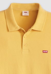 Cămașă polo galbenă dintr-un material texturat, cu guler clasic, plasă cu trei butoane și un mic logo roșu Levi's pe piept.