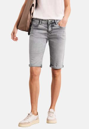 Femme portant un short en jean gris clair avec ourlet roulé, un t-shirt blanc, des baskets beiges et un sac à main beige porté à l'épaule.