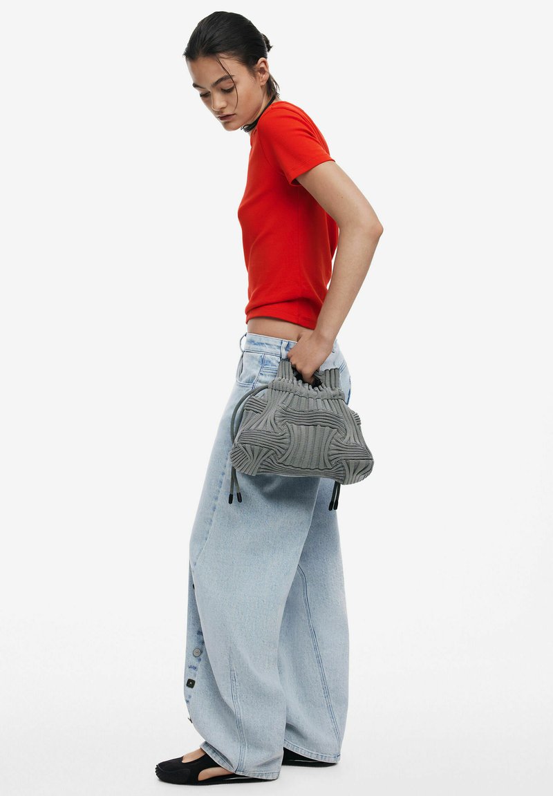 Grau geflochtene Tasche mit strukturierten Mustern, kombiniert mit einem roten T-Shirt und hellblauen weiten Jeans. Schwarze Ballerinas runden den lässigen Look ab.