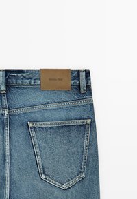 Tascone posteriore di jeans in denim blu con cuciture visibili e una toppa in pelle marrone con la scritta "Massimo Dutti" su sfondo bianco.