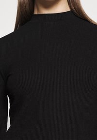 Schwarzes, geripptes Langarmoberteil mit hohem Kragen. Der Stoff wirkt strukturiert mit dezenten vertikalen Linien, die das figurbetonende Design unterstreichen.