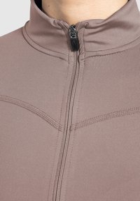 Lysbrun zip-up jakke med høj krave, der har en glat stoftekstur og kontrasterende syninger langs sømmene.