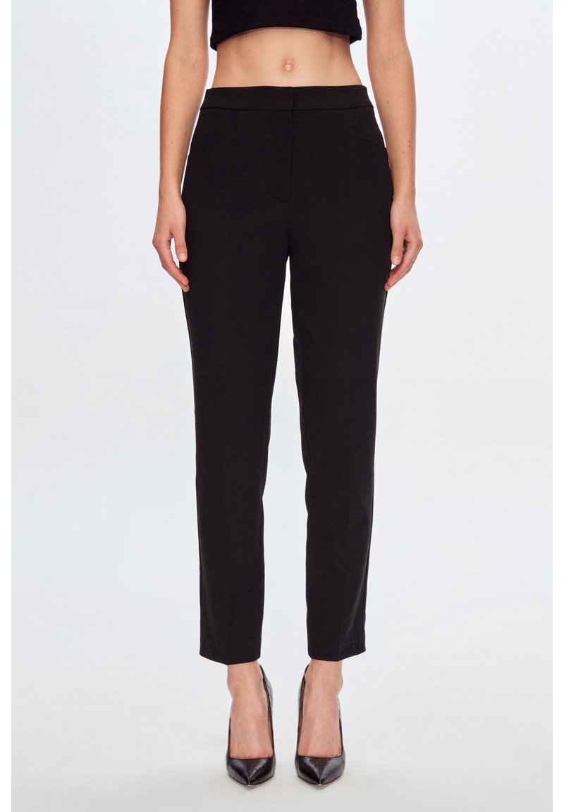 Femme portant un pantalon noir court, des talons aiguilles noirs et un haut court noir, vue de la taille aux pieds sur un fond blanc.