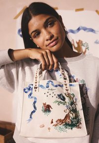 Torba na ramię z płótna w kremowym kolorze, z kolorowym haftem zwierząt, niebieskimi wstążkami i kwiatowymi ozdobami. Uchwyty ozdobione małymi dekoracjami.