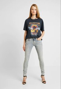 Jovem mulher em pé com a mão no bolso, vestindo uma t-shirt gráfica preta do Led Zeppelin, jeans skinny cinza claro e sandálias de salto pretas.