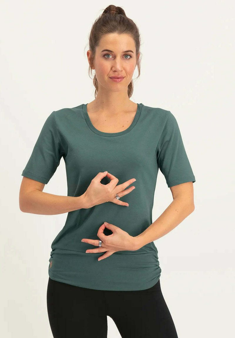 Türkisfarbenes T-Shirt mit kurzen Ärmeln und Rundhalsausschnitt. Weicher Stoff mit leichtem Stretch. Das Modell macht Handgesten, während es schwarze Hosen trägt.