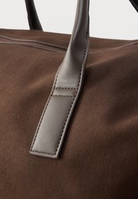 Καφέ σουέντ τσάντα χειρός με λουριά από λείο δέρμα και ορατά ραφές, που παρουσιάζει μαλακή υφή και δομημένο σχεδιασμό.